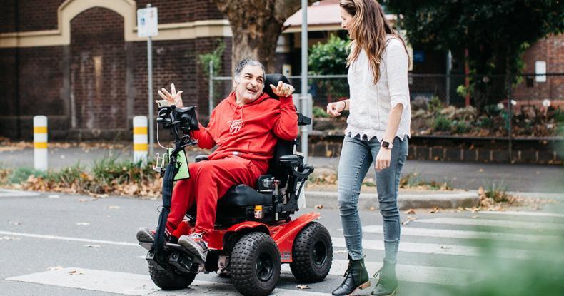 Man in mobility scooter wearing red tracksuit and woman wearing jeans and white shirt are crossing a pedestrian crossing and talking animatedly.