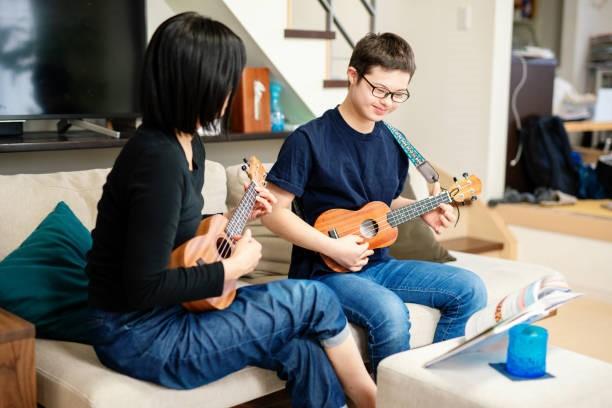 Boy with disability sitting on couch withd isability worker, both playing ukulele